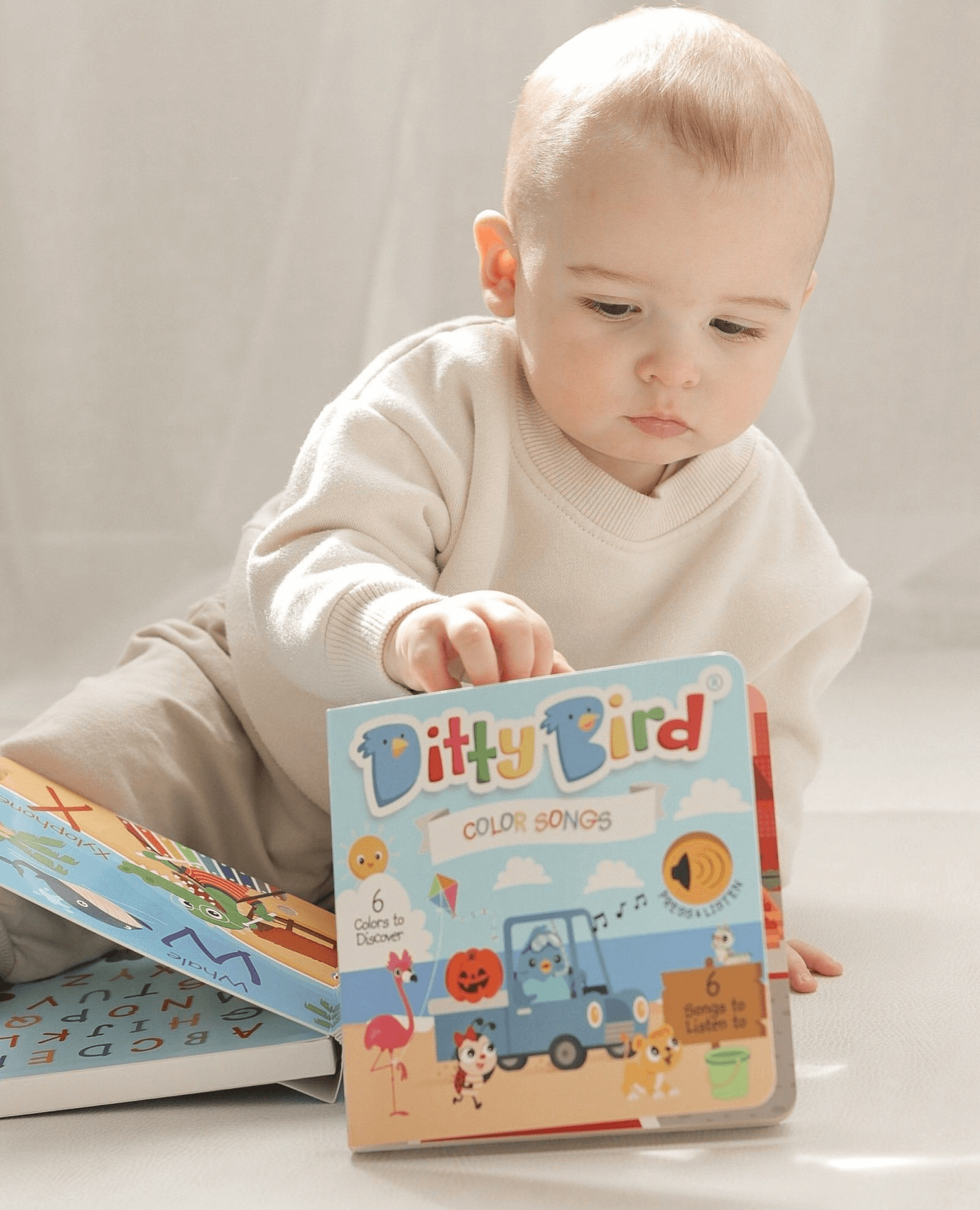 A baby in light clothing sits on the floor, holding a Ditty Bird Color Songs | AG10 model sound book by Ditty Bird—perfect for preschoolers learning colors. Another book lies open nearby against a softly lit, neutral background.