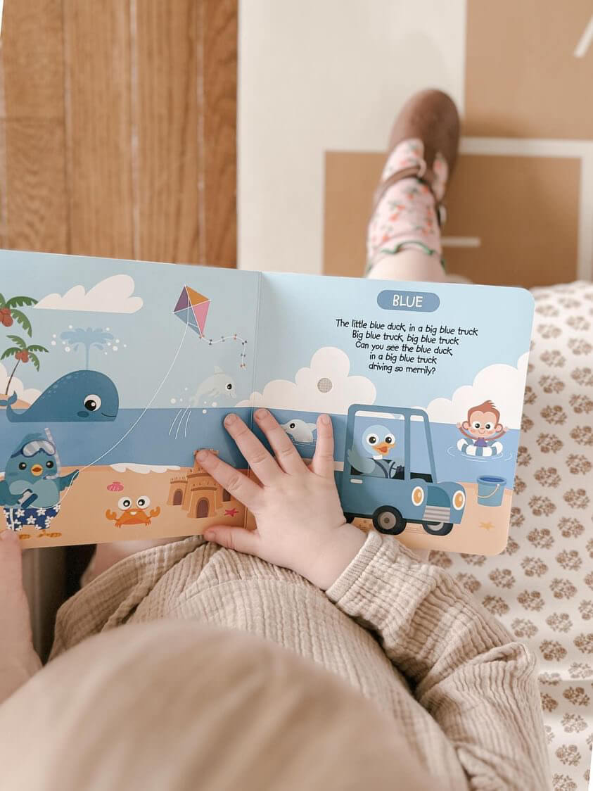 A child enjoys preschool fun with Ditty Bird's Color Songs | AG10 model on their lap, exploring colorful images like a blue whale, blue birds, a blue truck, and a monkey flying a kite as they learn colors.