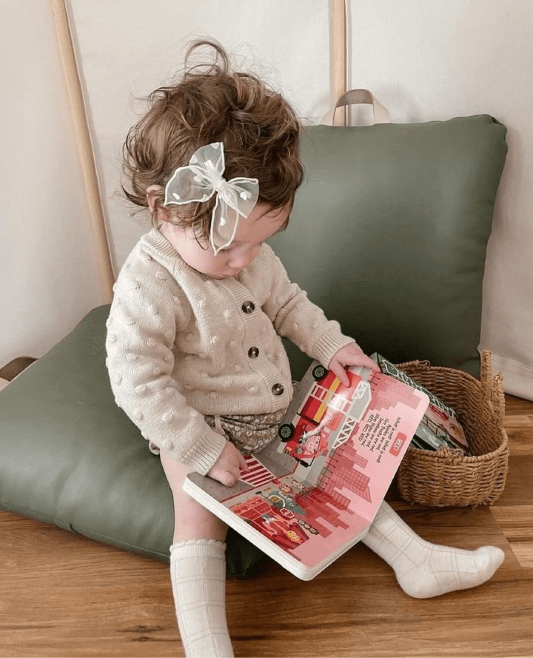 A toddler with curly hair and a sheer bow sits on a green cushion, wearing a cream sweater and socks, and explores the Ditty Bird Color Songs | AG10 model to learn colors. A wicker basket nearby adds to the preschool fun.