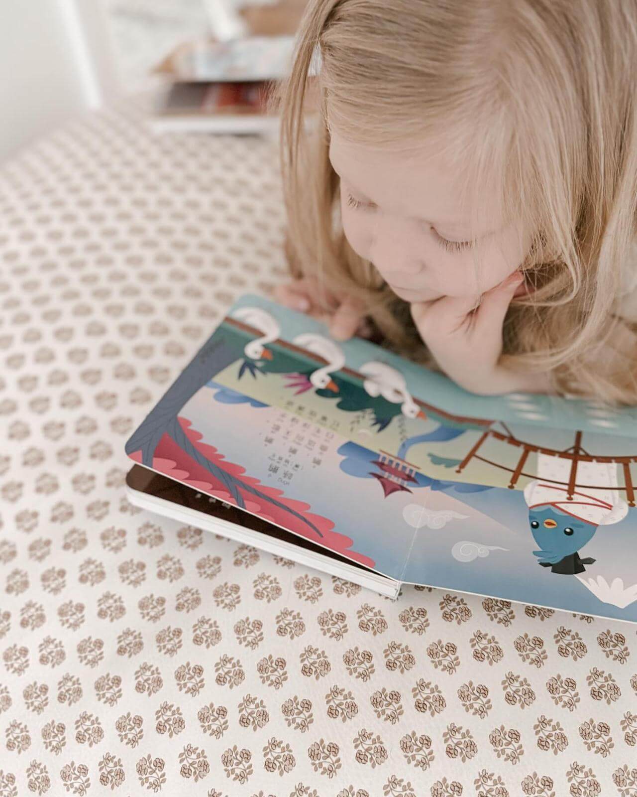 A young blonde girl lies on a patterned tablecloth, reading Ditty Bird’s "Singing Chinese Poems | AG10," an interactive children’s book featuring colorful drawings of birds, trees, and a bridge.