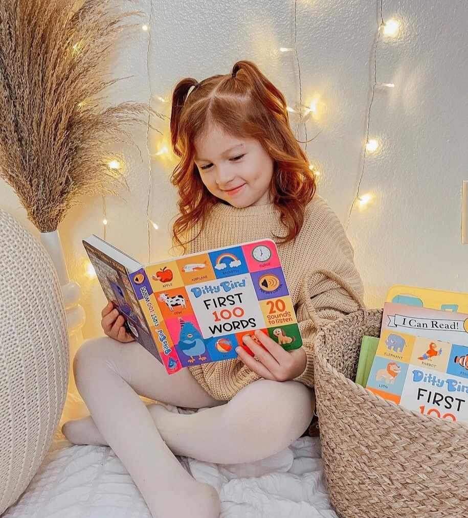 A young girl with red pigtails sits on a cozy blanket, smiling as she reads the Ditty Bird 100 Words Sound Book. Warm string lights and a basket of books nearby create an inviting space that fosters children's vocabulary learning.