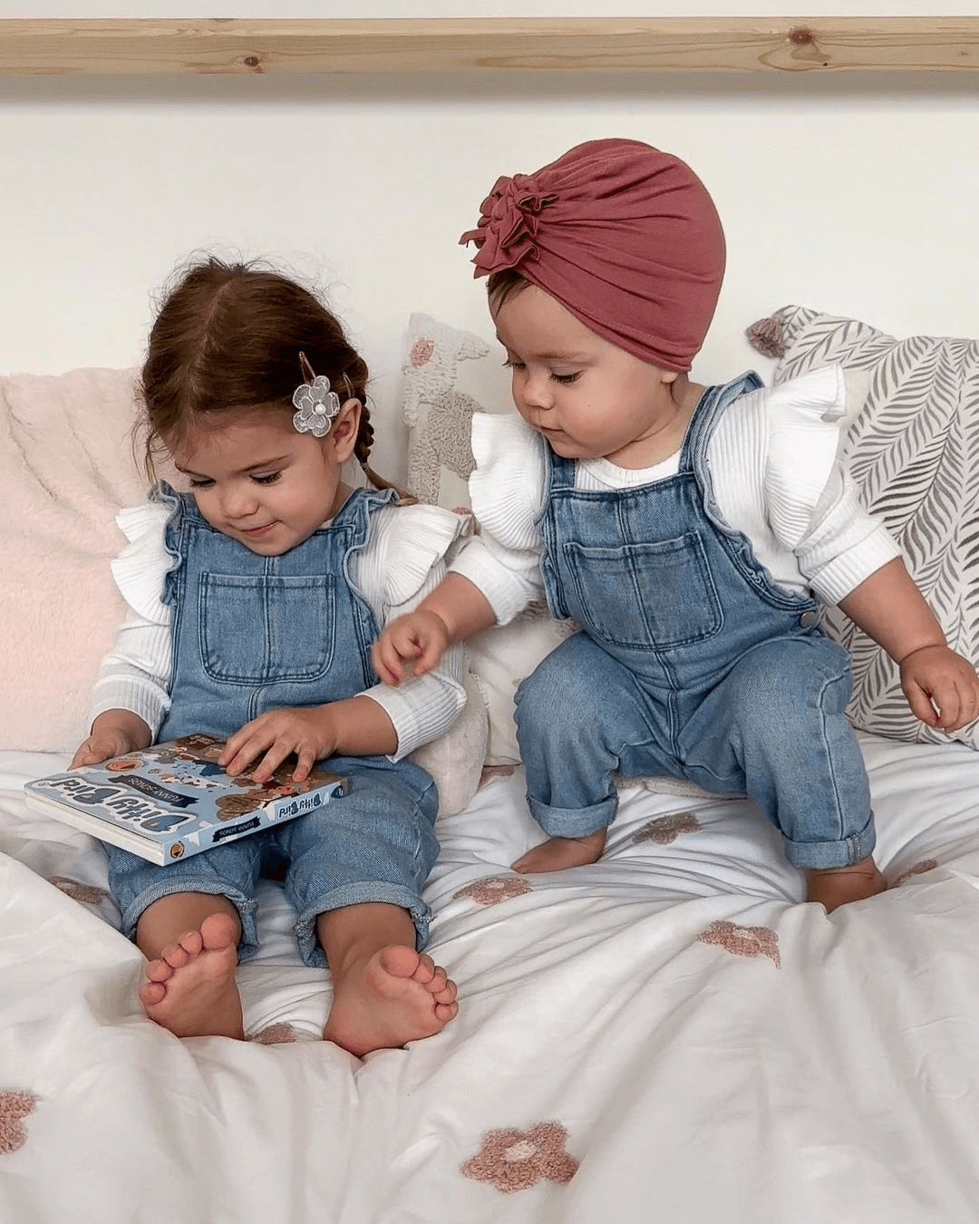 Two toddlers in matching denim overalls and white shirts sit on a bed. One reads the Ditty Bird "Funny Songs" music book while the other, in a pink headwrap, eagerly reaches for it among cozy cushions and a patterned blanket.