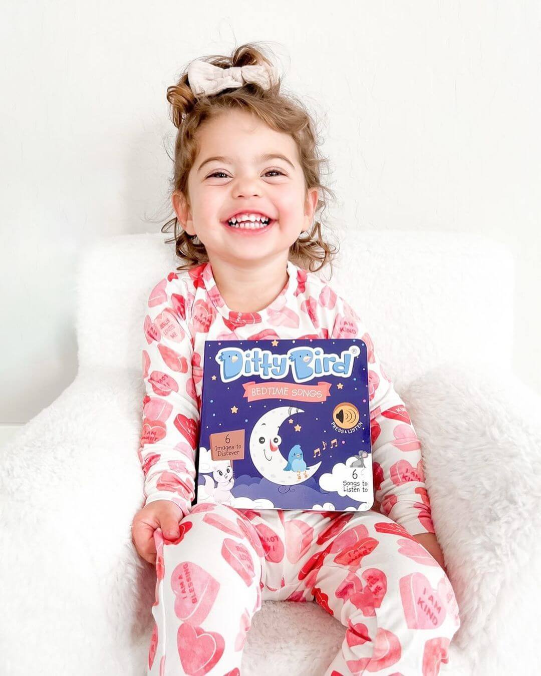 A young child in heart-patterned pajamas and a big hair bow sits on a white chair, smiling while holding the Ditty Bird Bedtime Songs book.