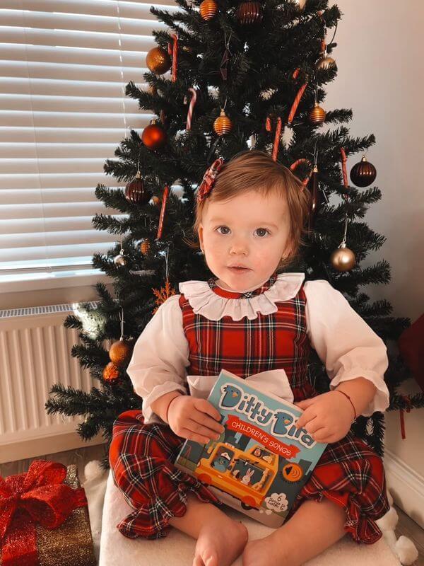 A young child in a red plaid dress sits by a Christmas tree, holding the Books “Children's Songs | USB-C rechargeable” interactive sound book, with wrapped presents nearby as she enjoys her favorite songs.