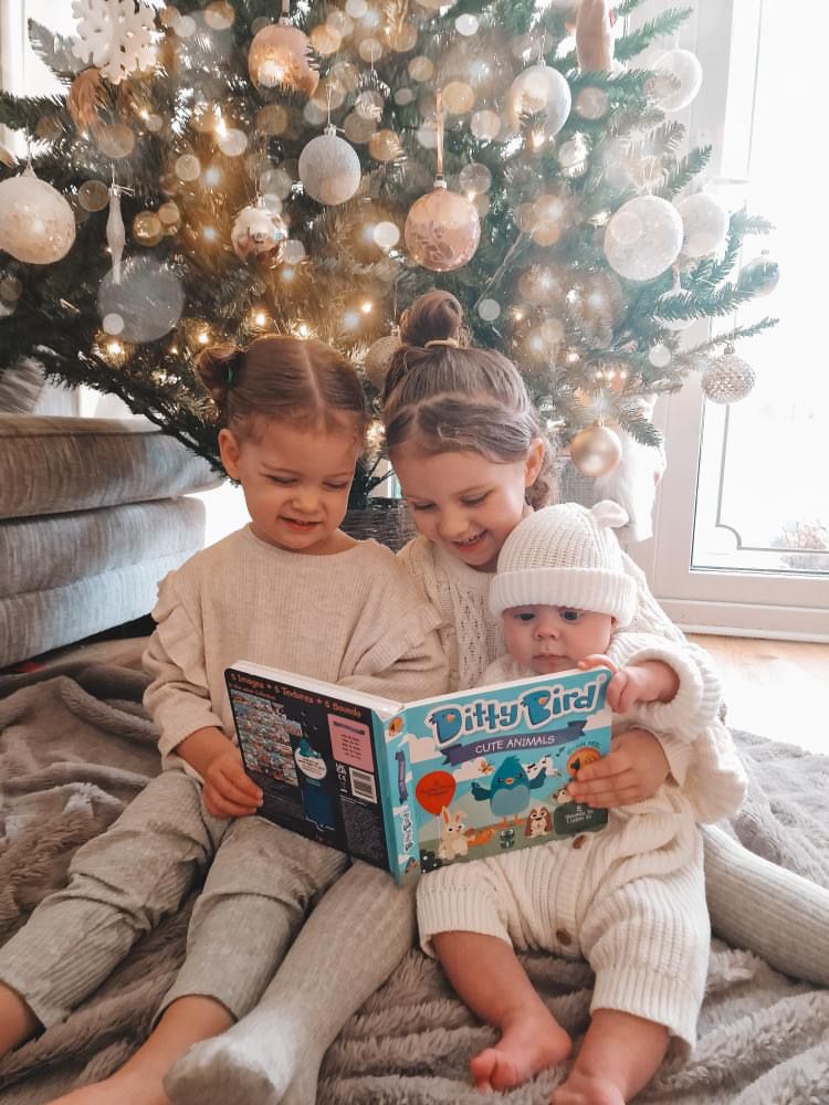 Three young children sit together on a rug in front of a decorated Christmas tree, smiling and reading Ditty Bird’s "Touch the Cute Animals" sound book. The youngest child wears a white knit outfit and hat. The book is USB-C rechargeable.