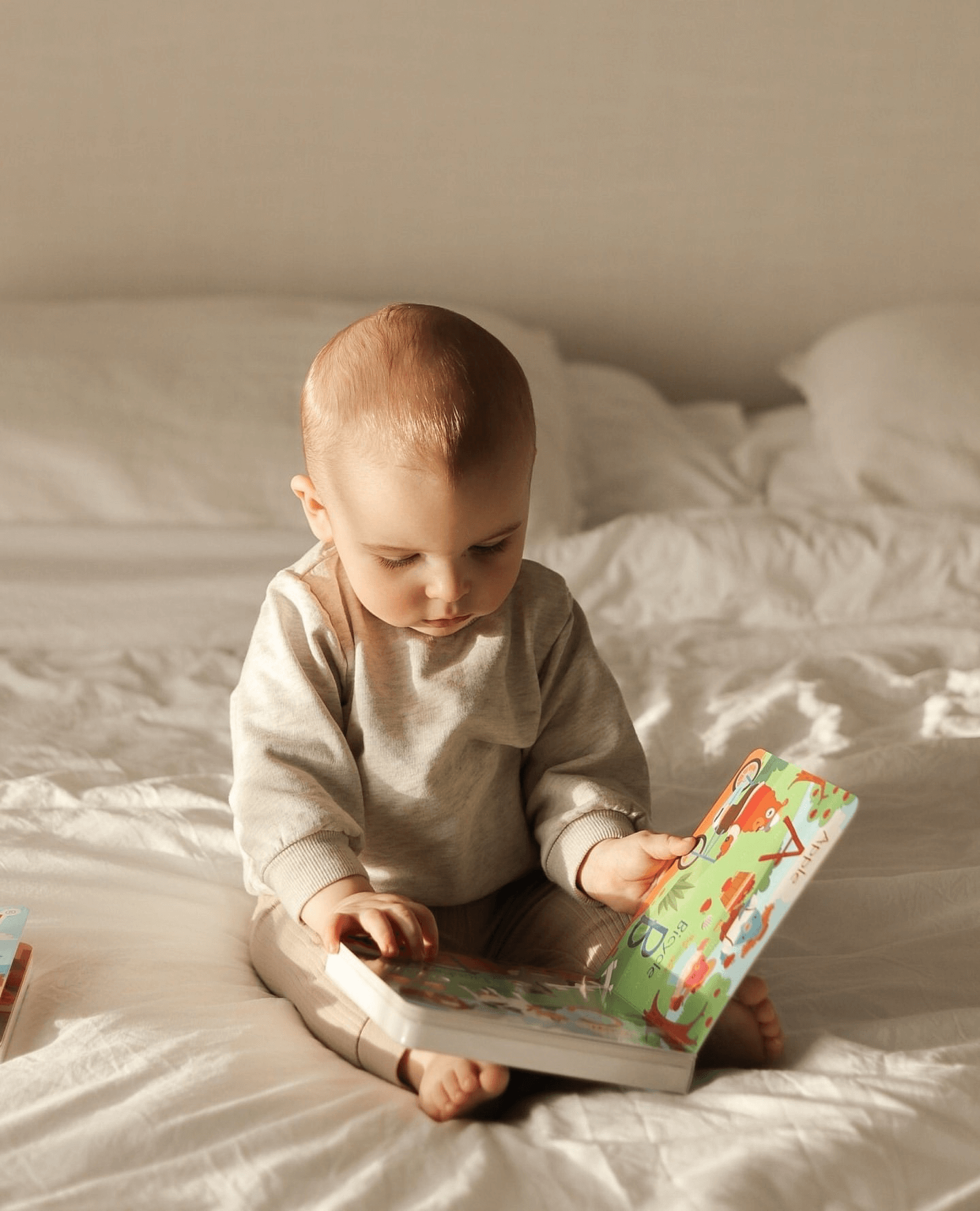 A baby sits on a bed in soft natural light, dressed in neutral tones, looking down at the Ditty Bird NEW! Spooky Halloween | AG10 book. White bedding and pillows create a peaceful background.
