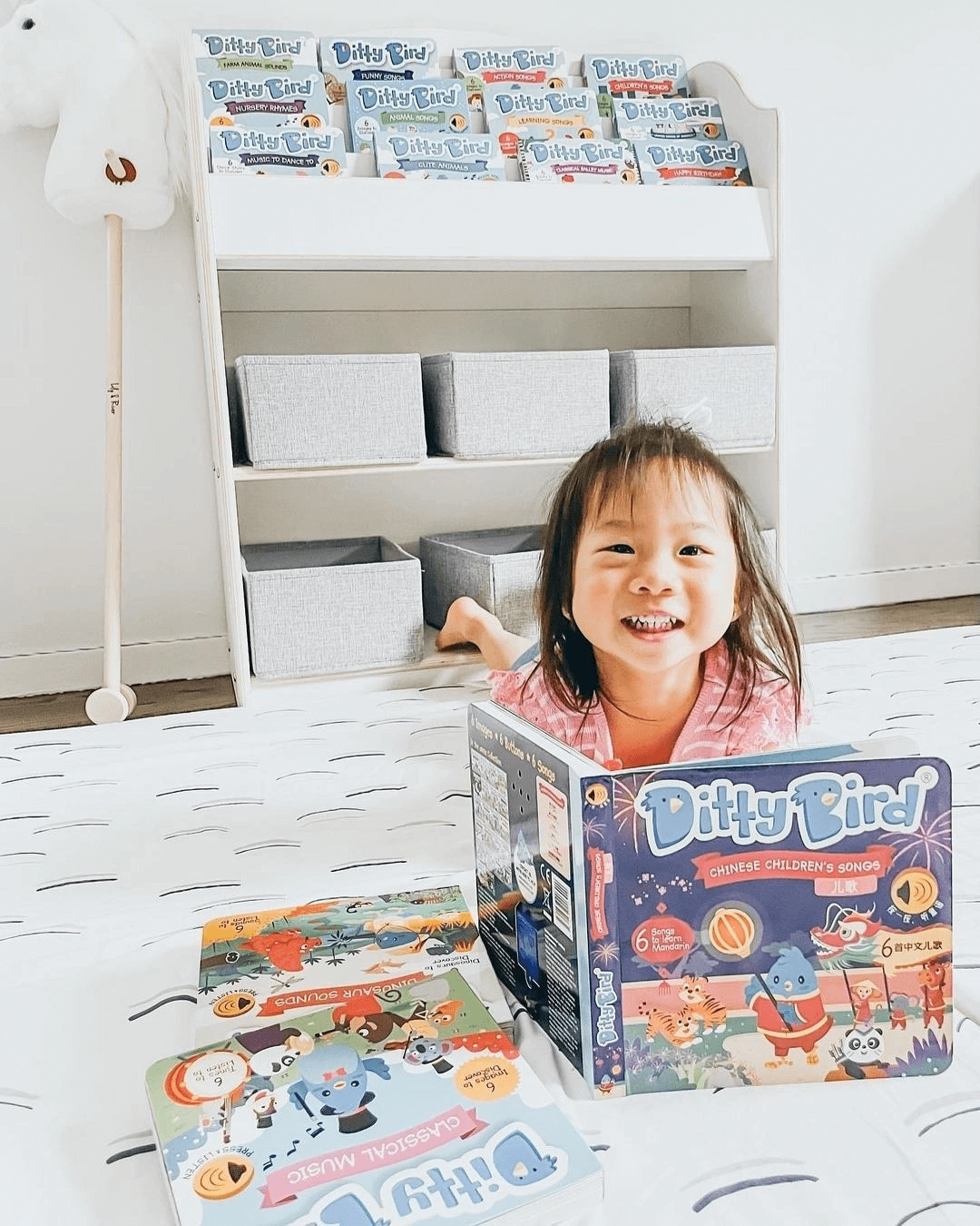 A young child smiles on a bed, holding the Books Chinese Songs Vol.1 sound book. Other Books titles lie nearby, with more displayed on a shelf in the bright, tidy room.
