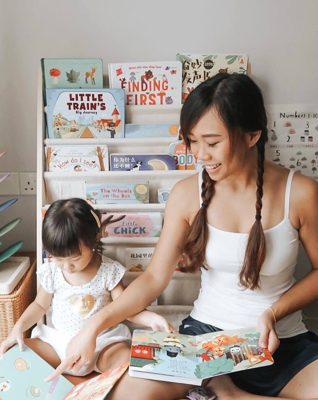 A woman and a young girl sit on the floor, smiling as they read children’s books together—including "Chinese Songs Vol.1" by Books—surrounded by a bookshelf filled with colorful picture books.