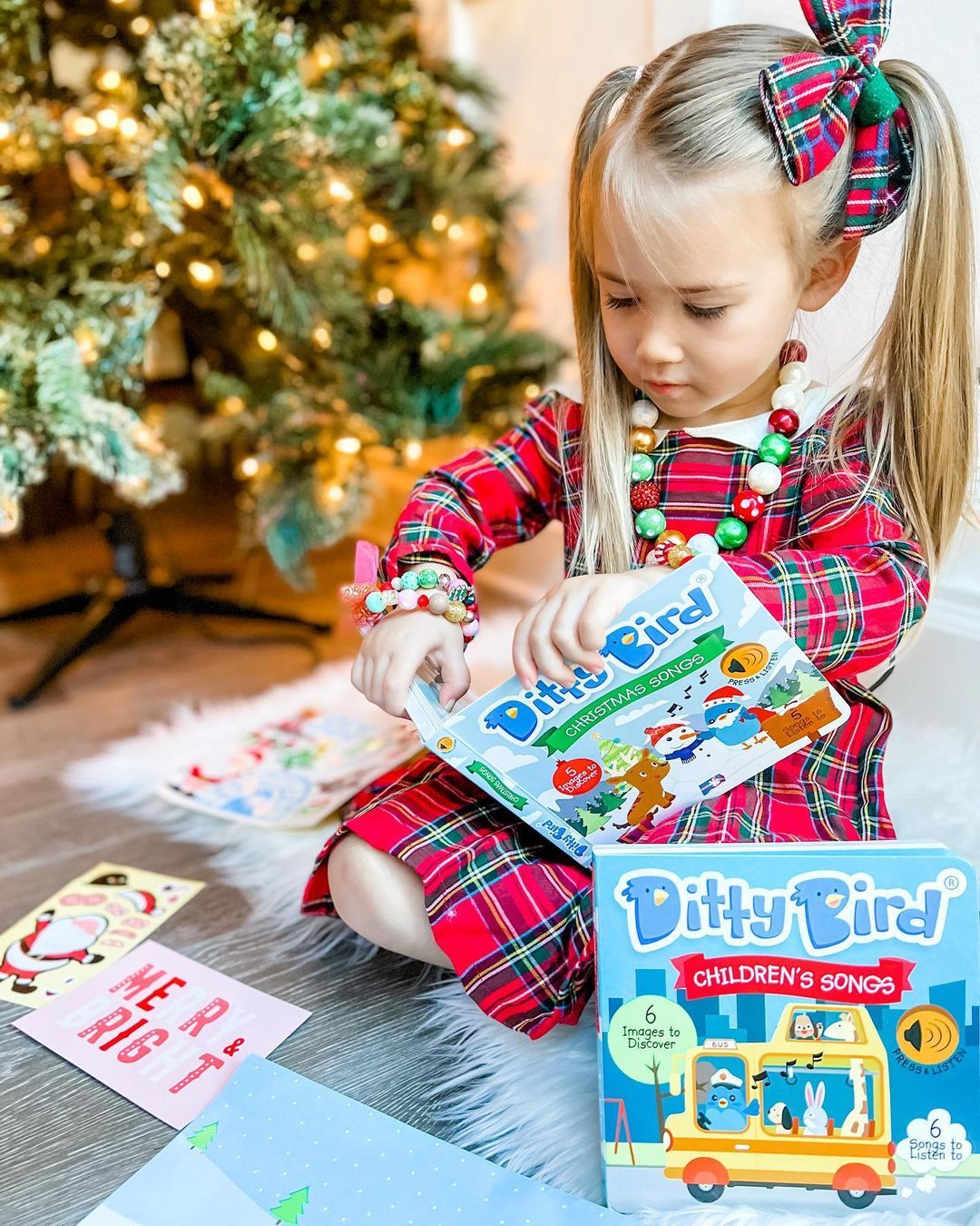A young girl in pigtails and festive bracelets sits by a Christmas tree, looking at the Books Children's Songs interactive sound book (USB-C rechargeable). Gifts and cards are scattered on the floor nearby.