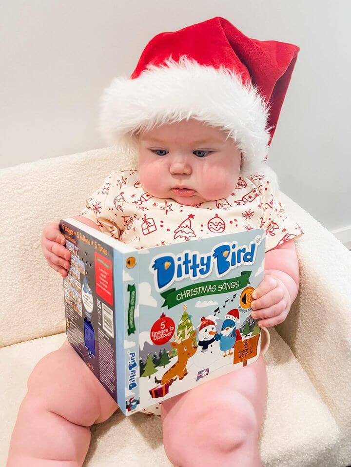 A baby in a Santa hat and festive pajamas sits on a chair, holding and looking at the Ditty Bird Christmas Songs book.