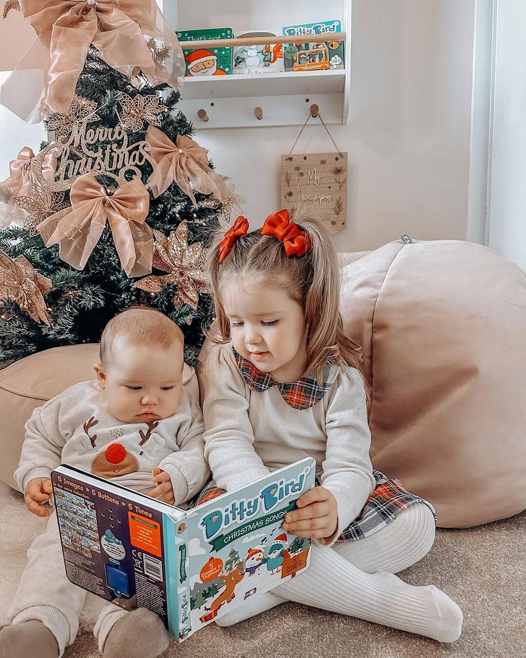 Two young children sit by a festive Christmas tree, reading Ditty Bird’s Christmas Songs together. The cozy scene, with bows, ornaments, and gifts beneath the tree, is perfect for sharing this musical holiday book.