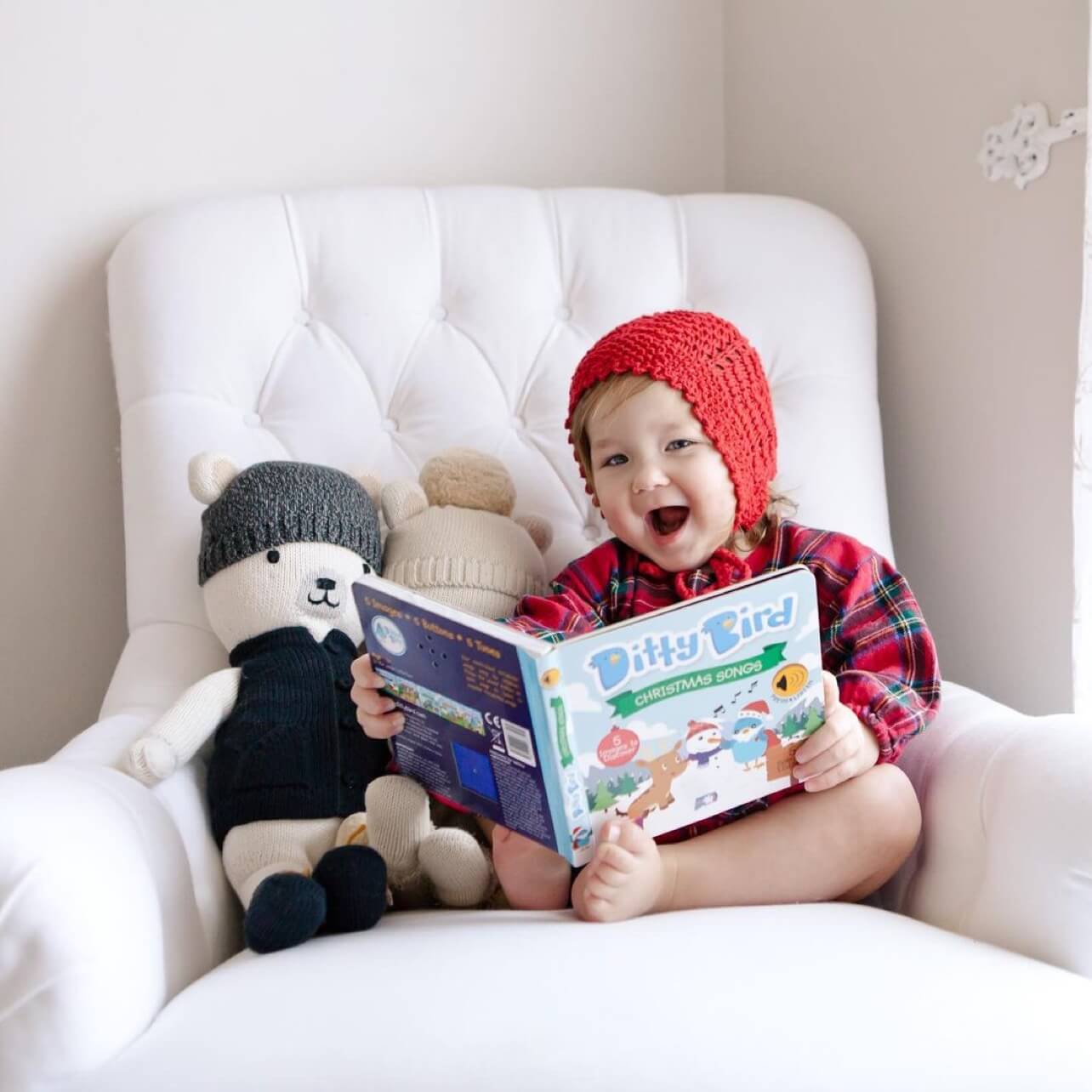 A young child in a red knit hat and plaid outfit sits on a white armchair, smiling and holding an open Ditty Bird Christmas Songs book, with two stuffed bears beside them.