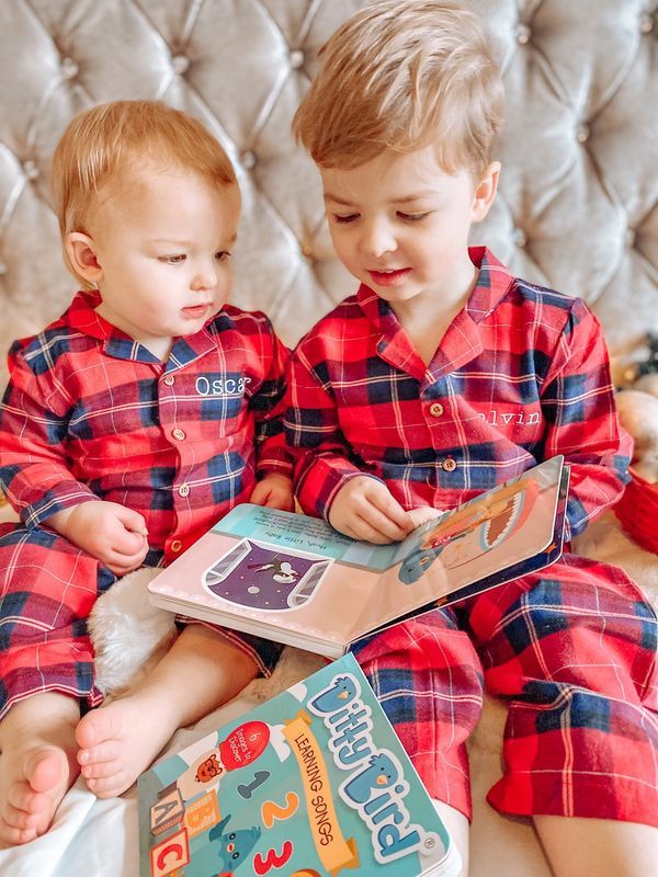 Two young children in matching red plaid pajamas with embroidered names sit together on a bed. The older child reads the Ditty Bird Learning Songs USB-C rechargeable interactive children's book as they both enjoy the story.