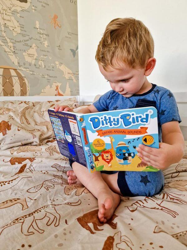 A young boy in blue pajamas sits cross-legged on animal-print sheets, reading Ditty Bird's Safari Animal Sounds book, which is filled with fascinating African animal sounds.