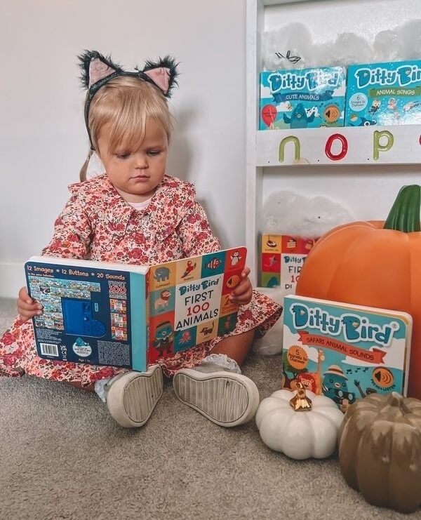 A young child in a floral dress and cat ear headband sits on the floor reading Ditty Bird's "Safari Animal Sounds" book, surrounded by other Ditty Bird books and decorative pumpkins near a bookshelf.