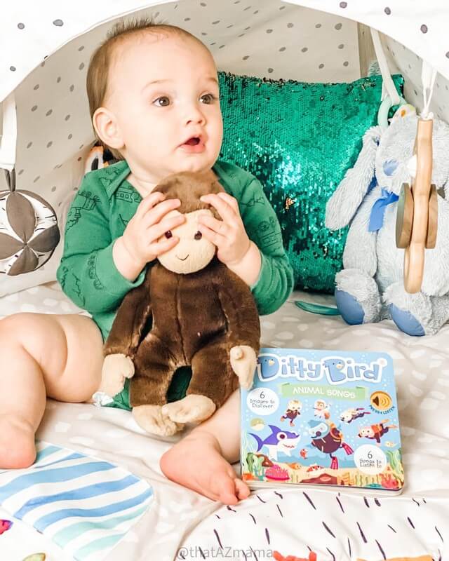 A baby sits on a soft play mat holding a brown stuffed monkey. Nearby are Ditty Bird’s Animal Songs interactive musical book, a blue stuffed bunny, and a shiny green sequin pillow in the background.