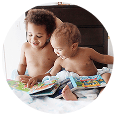 Two young children sit together on a cozy, well-lit bed, smiling as they explore interactive sound books. The scene captures the joy of early learning, with a dark dresser visible in the background.