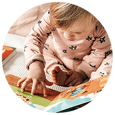 A baby wearing pink pajamas with black stars sits on a bed, reaching out and touching the pages of an interactive sound book—a perfect first birthday gift for curious little ones.