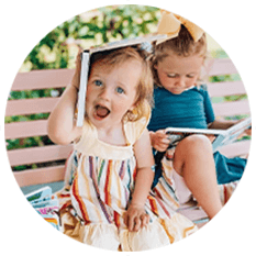 Two young children sit on a bench outdoors. One in a striped dress smiles playfully with a book on her head, while the other, wearing a blue top and yellow bow, looks down at an open book—capturing the joy of early learning.