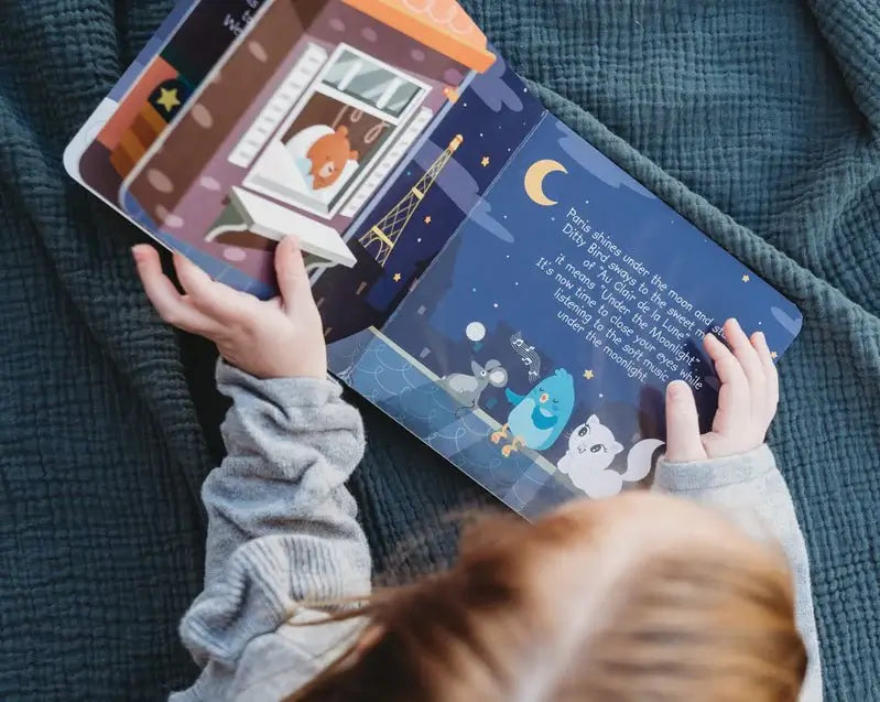 A child reads "Bedtime Songs" by Ditty Bird, a colorful picture book featuring animal characters under the moon and stars, like a gentle lullaby, while lounging on a blue blanket with text on the page.