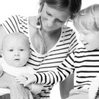 A woman holding a baby while a young child, perhaps searching for the perfect first birthday gift, reaches out to touch the baby’s hand. All three, dressed in striped shirts, are captured in this elegant black and white image.