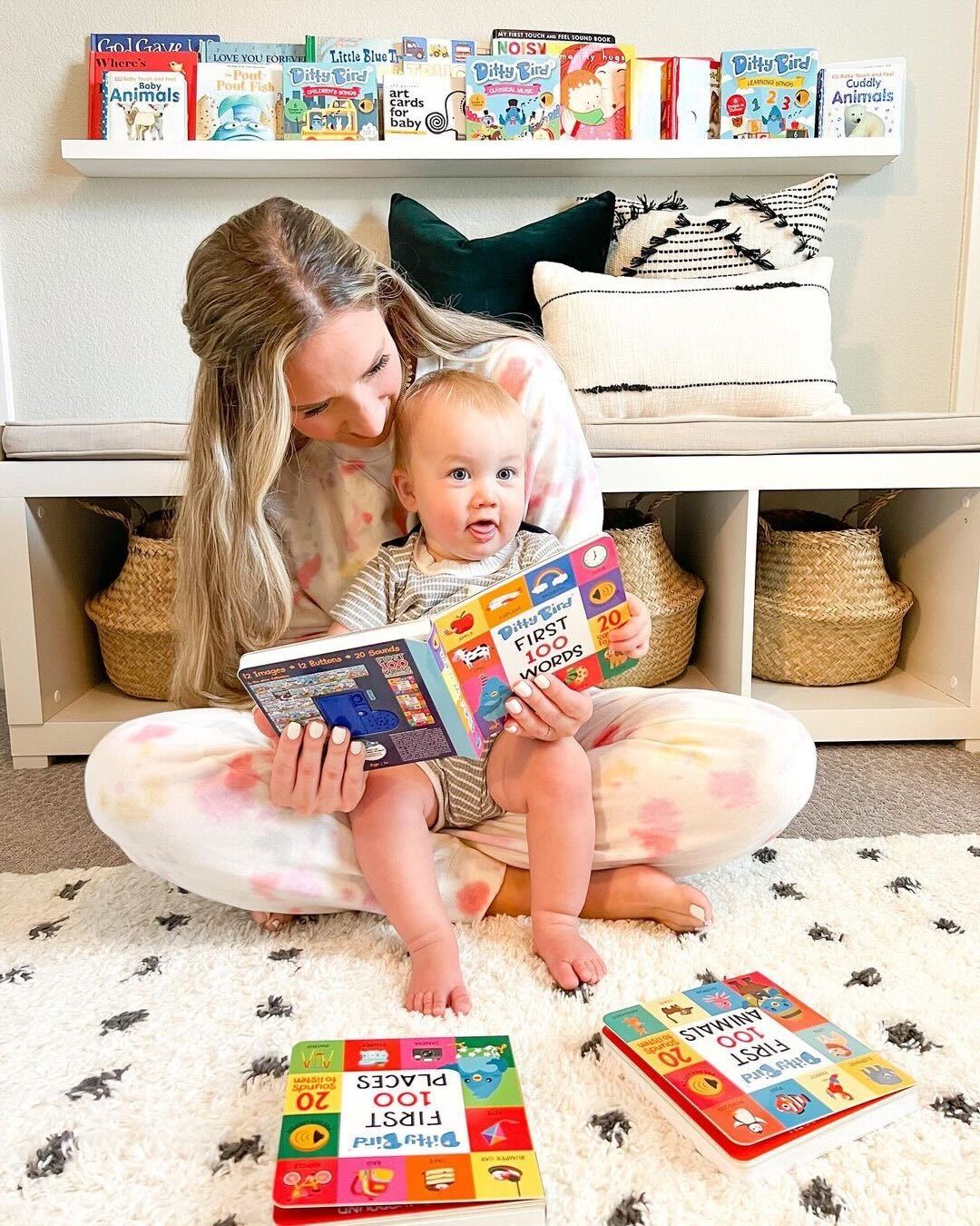 A woman sits on a rug with a baby in her lap, both reading the Ditty Bird 100 Animals Sound Book. Nearby are more interactive sound books, baskets, pillows, and a shelved educational safari book in a bright, cozy setting.