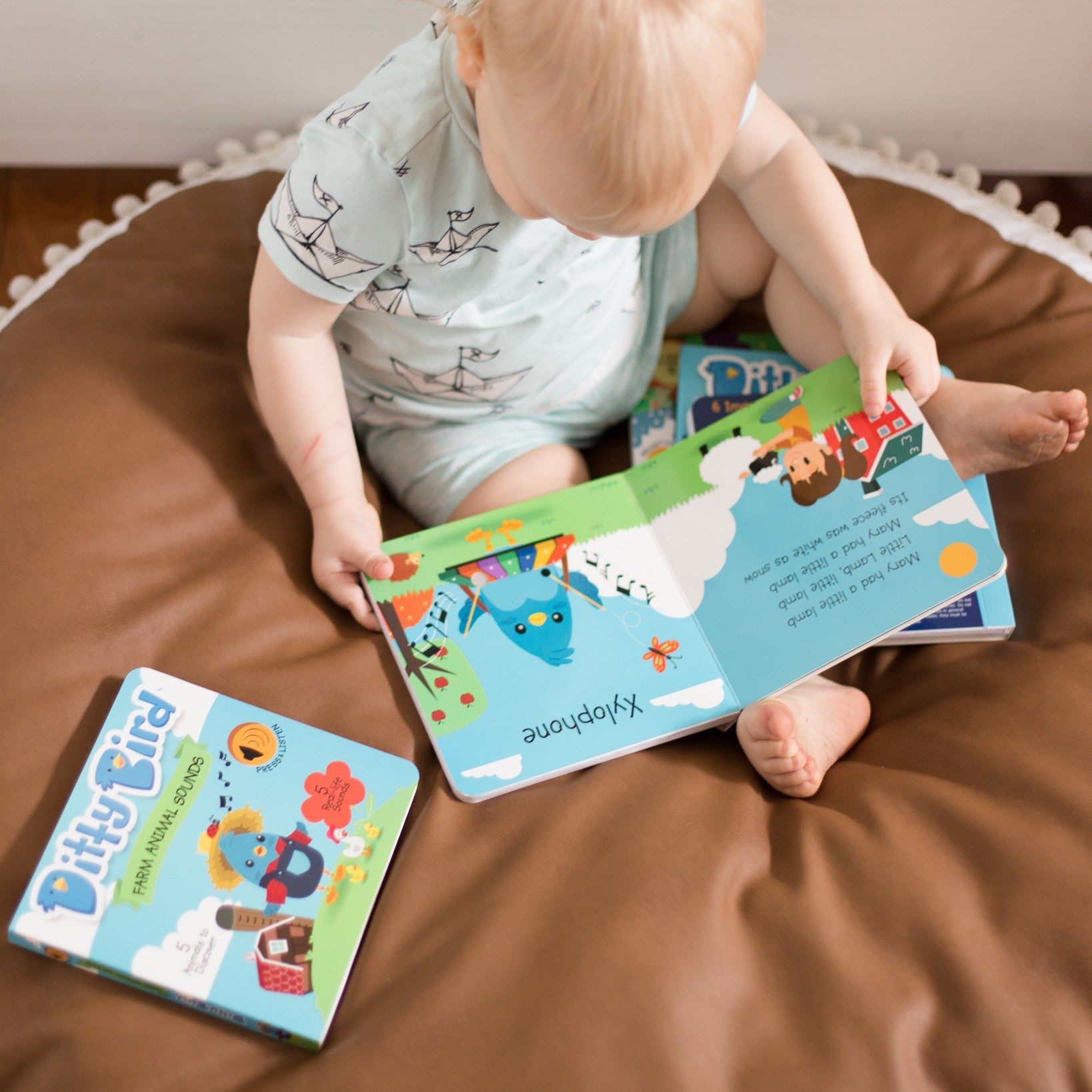 A toddler in a light blue outfit with bird prints enjoys the Ditty Bird Gift Set - Instrumental Songs on a brown bean bag, while another Ditty Bird book rests nearby—an adorable scene, perfect for a baby shower gift.