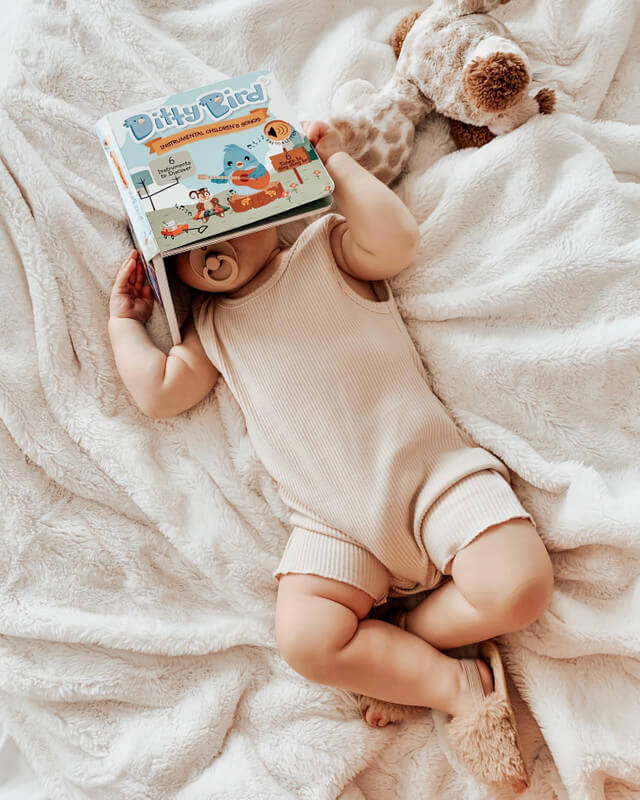 A baby in a beige romper lies on a soft, white blanket with a pacifier, holding Ditty Bird's Instrumental Songs sound book over their face. A plush giraffe toy nearby invites playful musical exploration.