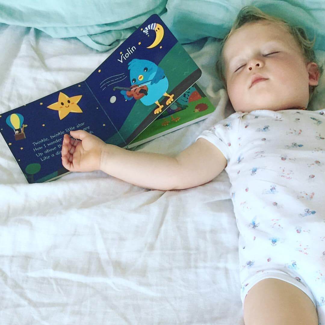 A young child in a white onesie sleeps on a bed beside the Ditty Bird "Instrumental Songs" book, which is open to a page with a blue bird playing violin under the moon and stars, inspiring dreams of music.
