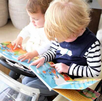 Two young children sit together, each reading a Ditty Bird "Instrumental Songs" sound book. One wears a striped shirt and navy vest, the other is in white. Both are engaged in musical exploration near a toy steering wheel.