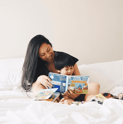 A woman and a young child smile as they sit on a bed, happily reading Ditty Bird's Instrumental Songs, a colorful children's sound book that makes musical exploration fun and engaging.