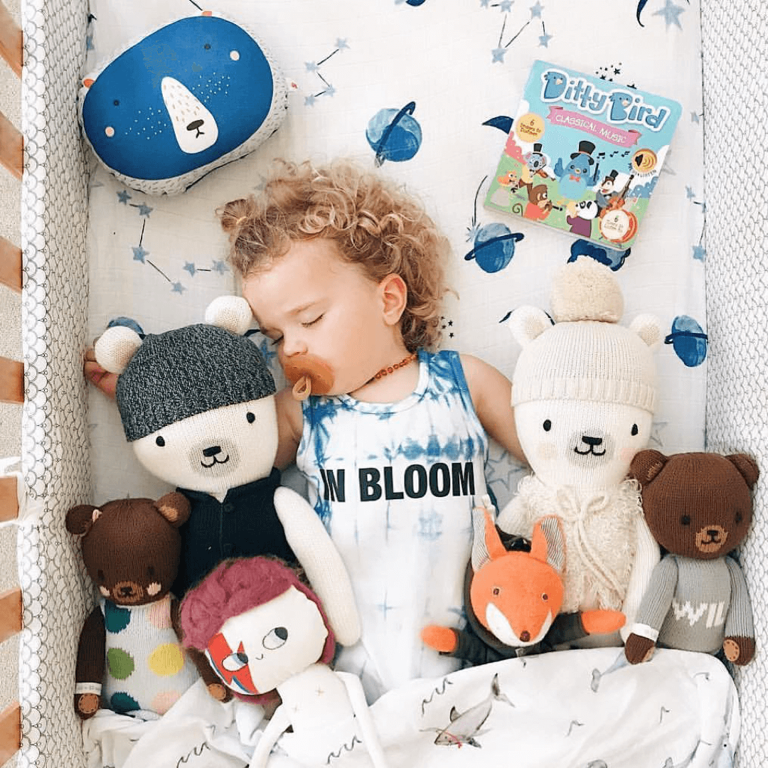 A young child with curly hair naps in a crib with stuffed animals, holding a pacifier and wearing an ON BLOOM tank top, while resting beside Ditty Bird’s "Classical Music" sound book to support cognitive development.
