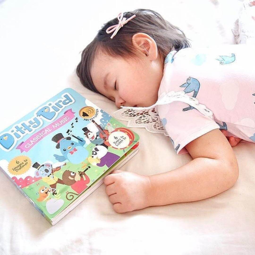 A young child with short dark hair, wearing a pink outfit with blue birds, sleeps on a bed next to Ditty Bird’s "Classical Music" sound book—an ideal way to encourage cognitive growth and introduce music to kids.