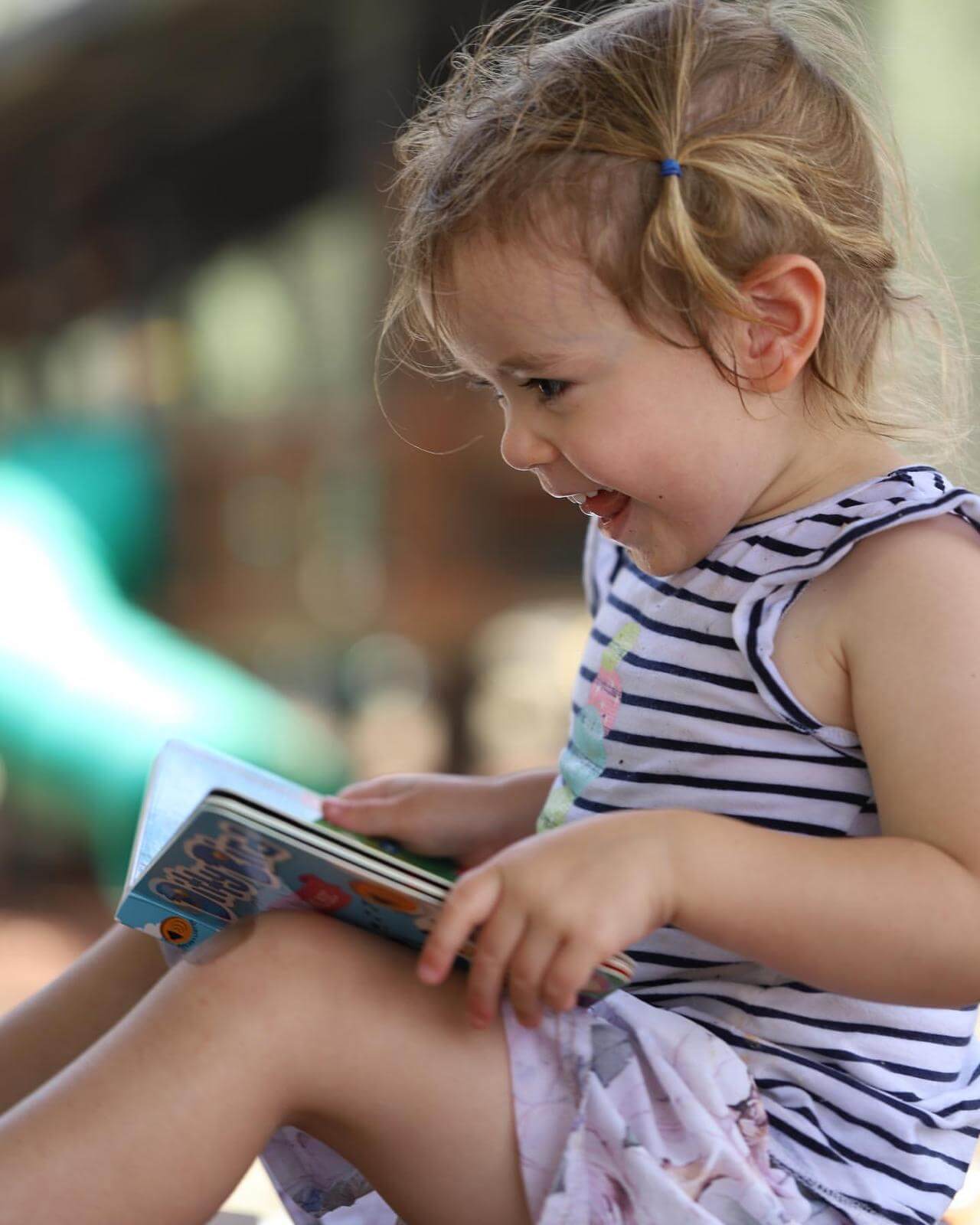 A young girl with light brown hair in a blue clip, wearing a striped dress, smiles while reading the Ditty Bird "Classical Music" sound book outdoors. A green slide and blurred background suggest she's at a playground.