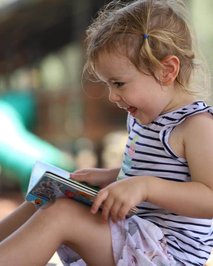 A young girl with light brown hair in a blue clip, wearing a striped dress, smiles while reading the Ditty Bird "Classical Music" sound book outdoors. A green slide and blurred background suggest she's at a playground.