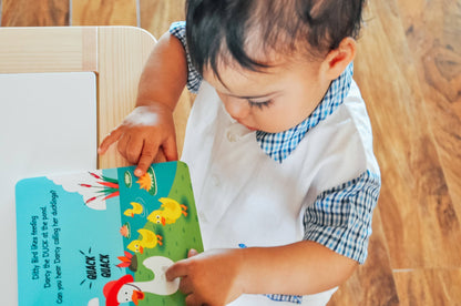 A toddler in a white shirt with blue trim looks down and points at the Ditty Bird "Farm Animal Sounds" book featuring colorful farm animal illustrations on a wooden floor.