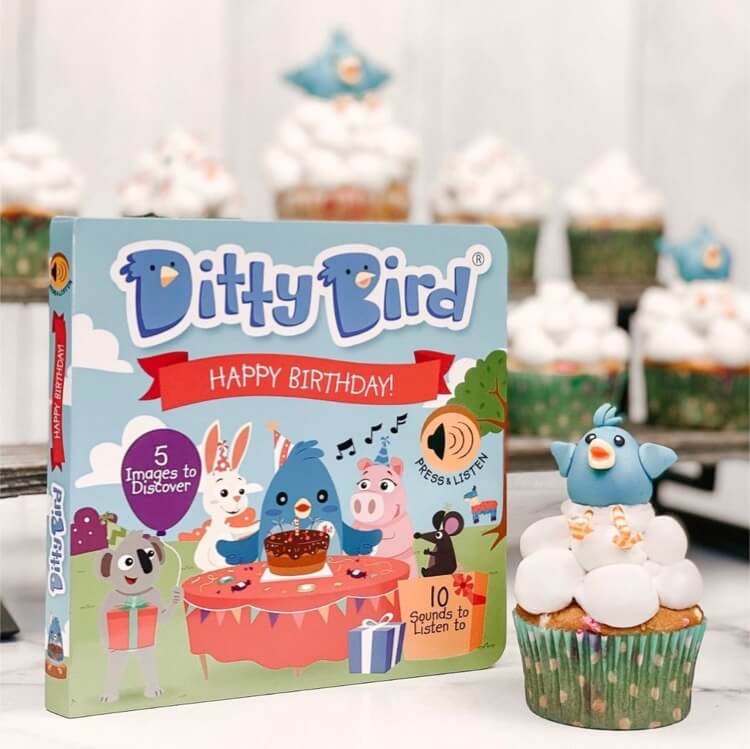 The Ditty Bird "Happy Birthday" musical book sits next to a cupcake with a blue bird topper and white icing. More bird-topped cupcakes appear in the background—a charming birthday gift for kids.