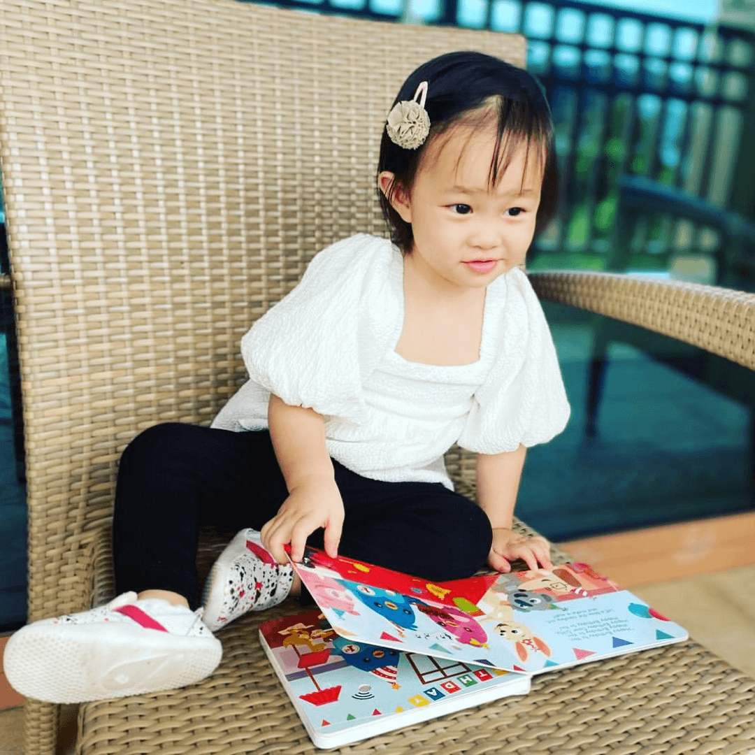 A young child with a hairclip sits on a wicker chair outdoors, smiling at an open Ditty Bird "Happy Birthday" musical book in their lap. The child wears a white top, black pants, and white shoes.