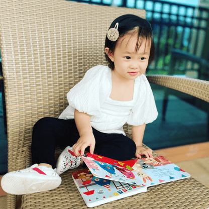 A young child with a hairclip sits on a wicker chair outdoors, smiling at an open Ditty Bird "Happy Birthday" musical book in their lap. The child wears a white top, black pants, and white shoes.