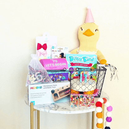 A small round table holds Ditty Bird's "Happy Birthday" basket for kids—with toys, books, a plush duck in a pink party hat, cake, decorations, a pom-pom garland, and the interactive Ditty Bird "Happy Birthday" book for extra fun.