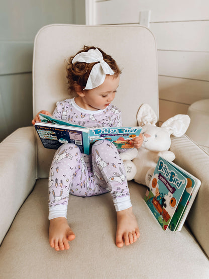 A young child in pajamas and a white headband sits on a beige armchair reading Ditty Bird's "Music to Dance to," with a plush white bunny and another book beside her, ready to enjoy lively tunes and fun movement.