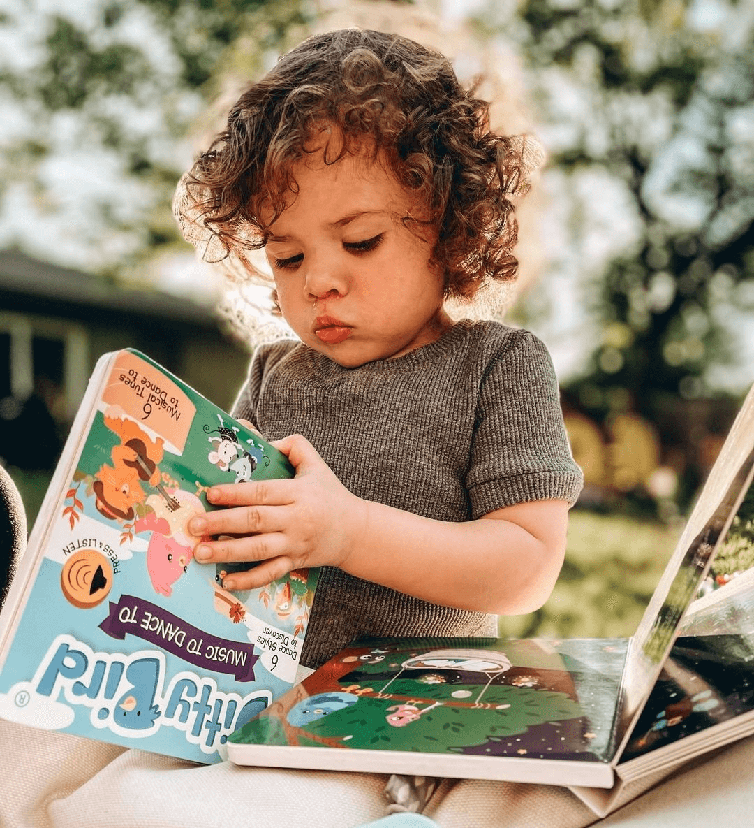 A young child with curly hair sits outdoors on a blanket, intently exploring the colorful Ditty Bird "Music to Dance to" musical book. Another open book rests on their lap, with green trees and sunlight in the background.