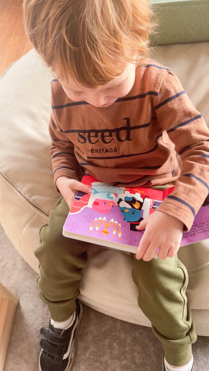 A young child in a brown striped shirt, olive green pants, and black sneakers sits on a beige chair, engaging with the Ditty Bird "Music to Dance to" musical book and touching its colorful pages.