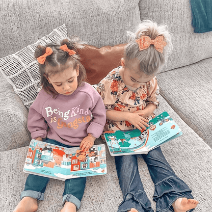 Two young girls sit on a gray couch reading Ditty Bird’s “Music to Dance to,” a musical book with colorful illustrations. One wears a purple “Being Kind is Gangster” sweatshirt; the other wears a floral top and jeans.