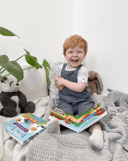 A young child with light red hair smiles happily while sitting on a cozy gray blanket with Ditty Bird’s Dinosaur Sounds book in their lap. Stuffed animals, a plant, and another book are nearby in the bright, cheerful setting.