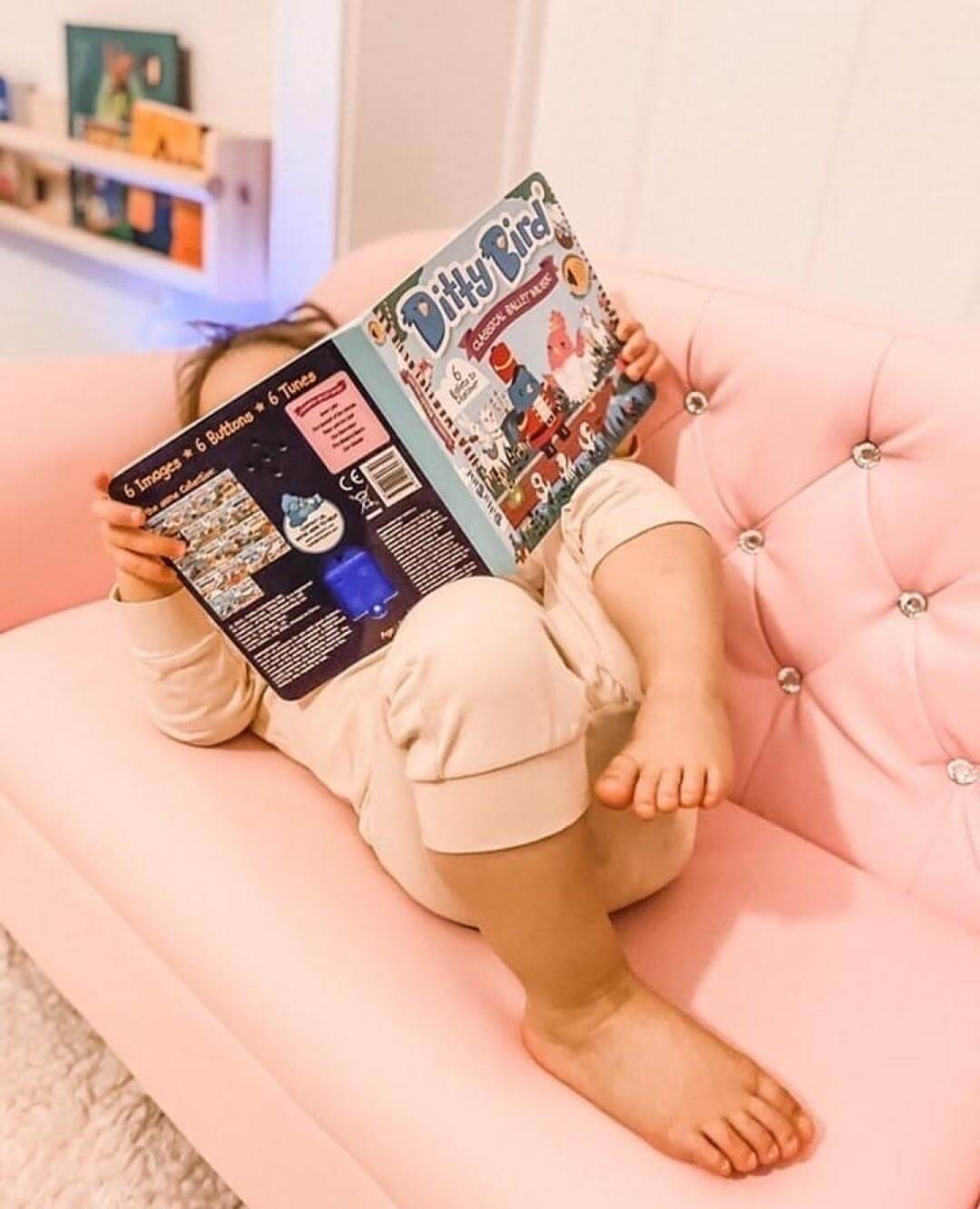 A young child relaxes on a light pink, tufted couch with legs up, face hidden behind the Ditty Bird Classical Ballet Music book as they explore colorful pages and classical ballet melodies.