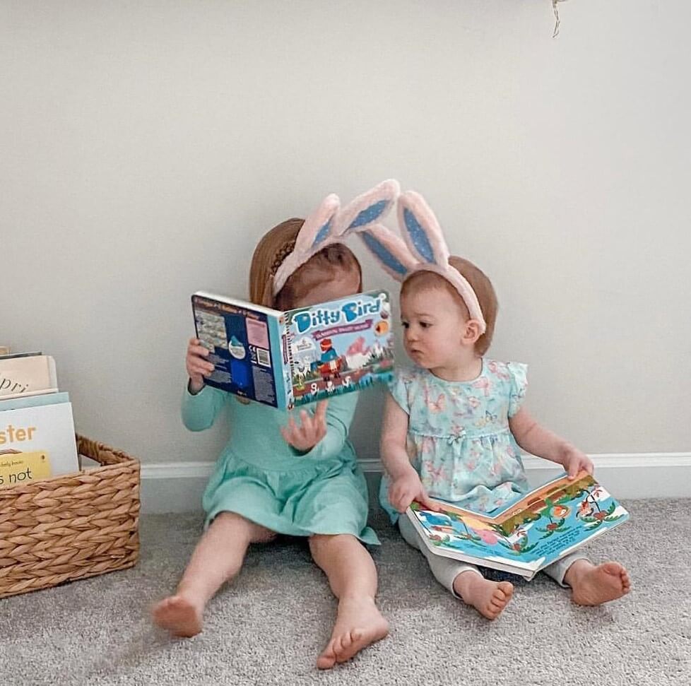 Two young children in pastel dresses sit on the floor reading books. One, wearing bunny ears, holds up Ditty Bird’s Classical Ballet Music book while the other looks at her own. A basket of more books is nearby.