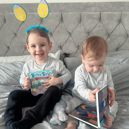 Two preschoolers in matching grey outfits sit on a bed with grey bedding. The older child, wearing blue Easter bunny ears, smiles while holding Ditty Bird’s interactive book “Career Songs.” The younger one looks down, focused on another book.