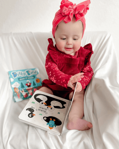 A baby in a red outfit and headband sits on a white blanket, smiling as she turns the pages of Ditty Bird's Black & White Animals high-contrast sound book. Another colorful children’s book lies beside her.