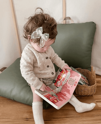 A toddler with curly hair and a sheer bow sits on a green cushion, wearing a cream sweater and socks, and explores the Ditty Bird Color Songs | AG10 model to learn colors. A wicker basket nearby adds to the preschool fun.